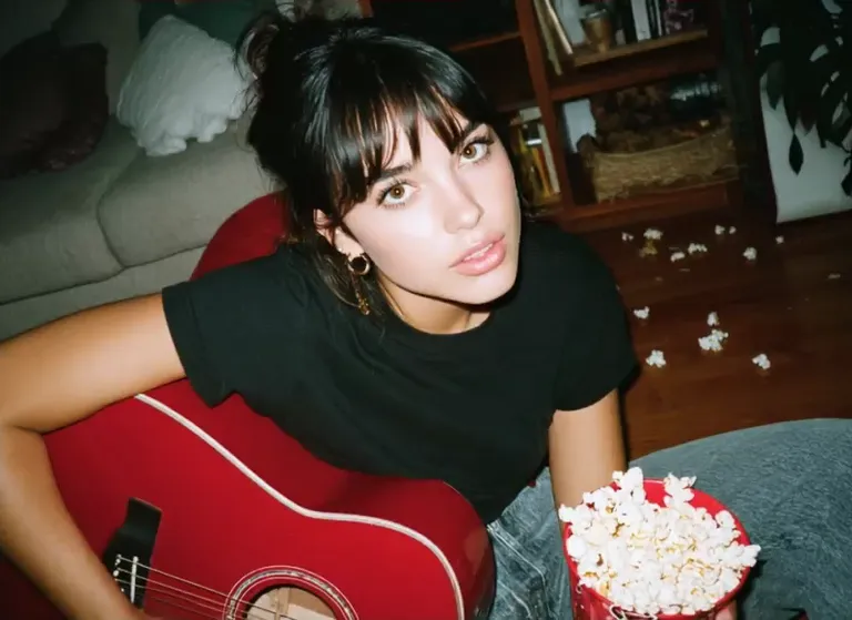 A young woman with a topknot sits by a gray couch holding a red acoustic guitar with a bowl of popcorn beside her, a real-person input photo for an AI ahegao video