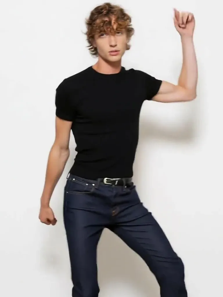 A curly-haired man in a black tee and jeans dances with one arm raised in a white studio, a real-person input photo for an AI twerk video